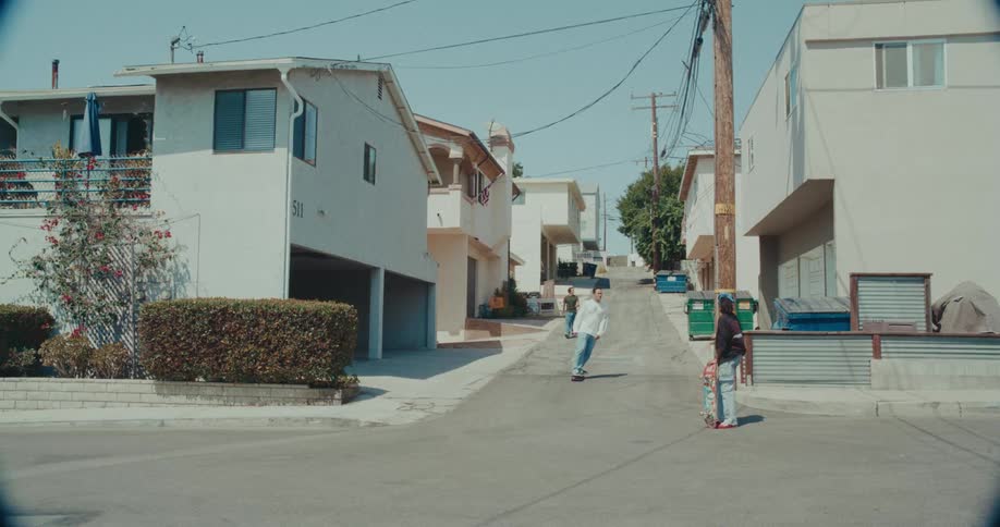 Female skateboarder holds her skateboard while standing in an intersection and watching a man skateboard past her
