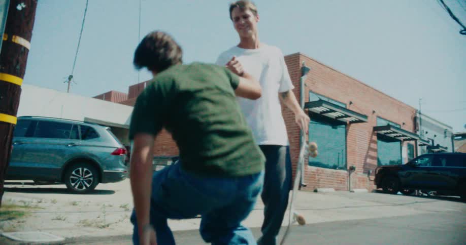 A man in a white tshirt carrying a skateboard helps another man in a green tshirt get up from the asphalt street near a brick building on a sunny day