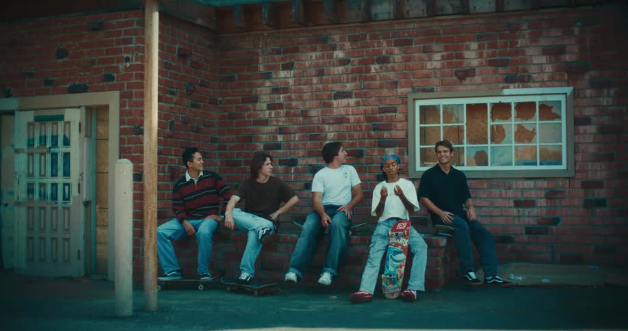 Five young skaters sit on a brick ledge outside a building with a broken window talking and laughing together
