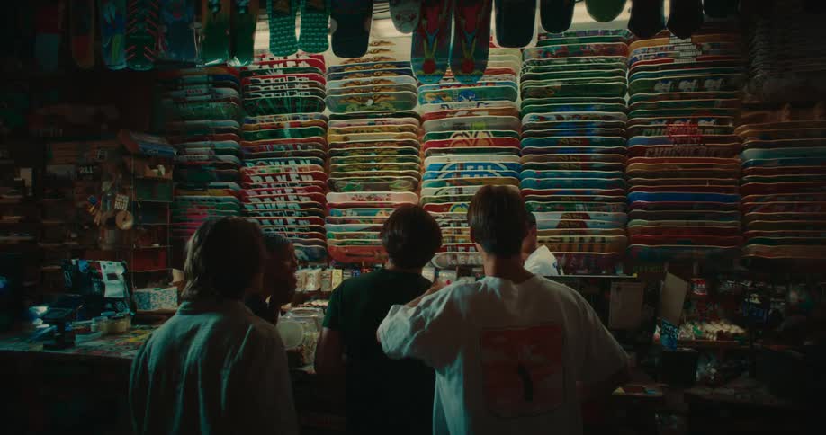Man working in a skateboard shop puts a skateboard on the stack behind the counter as a small group of young people come in to look at skateboards