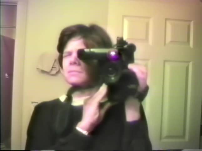 A woman with short dark hair holds a large black video camera to her eye while looking toward a mirror in a room with a white door