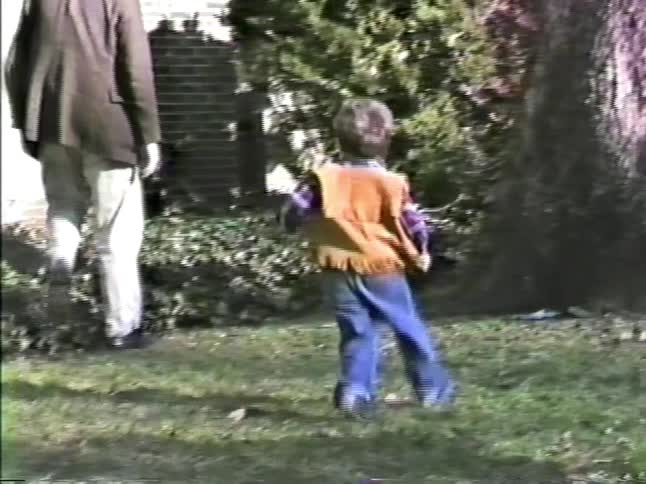 A man in a brown blazer and tan pants walks with two young boys in a grassy yard in front of a brick house with white shutters on a sunny day
