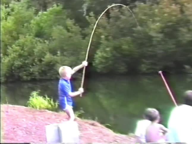 A young boy in a blue shirt stands on a grassy bank and pulls a fish out of a pond with a long rod while a man and two other children sit nearby and watch him