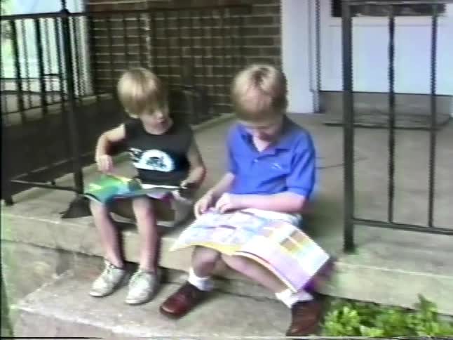 Two young boys sit in front of a house while reading through a magazine