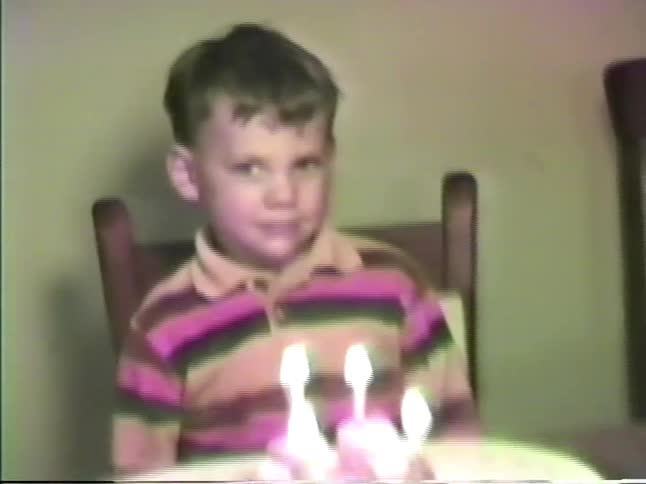 A young boy in a striped polo shirt sits at a wooden table and blows out white candles on a small chocolate cake during a birthday celebration