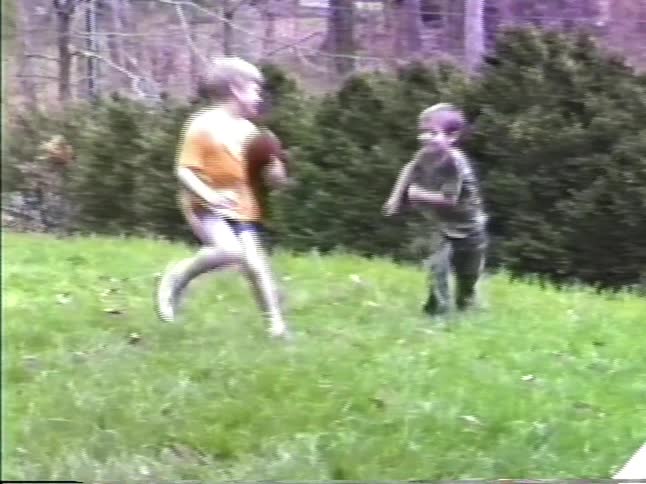 Two young boys in a backyard play catch with a football on a grassy hill and a third boy appears at the end of the shot