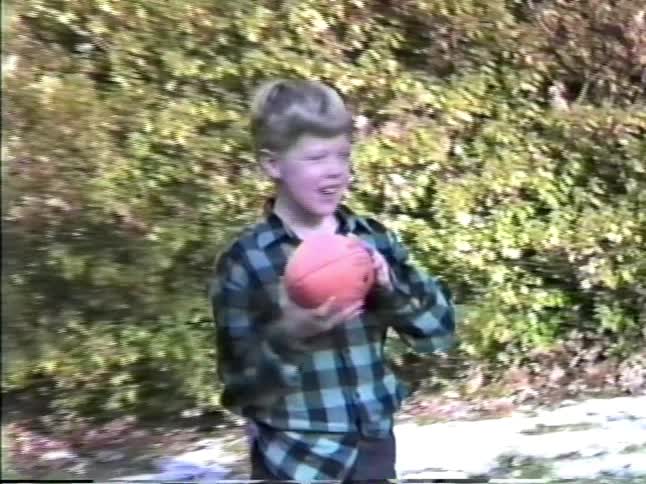 A young boy wearing a blue and black flannel shirt throws a red ball while playing with other children in a snowy yard lined with green bushes on a sunny day