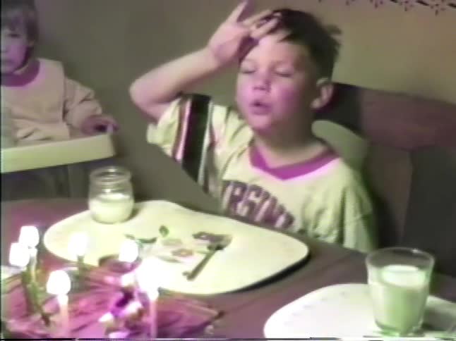 A family is sitting at a table as the mother lights candles on a birthday cake and one boy blows them out