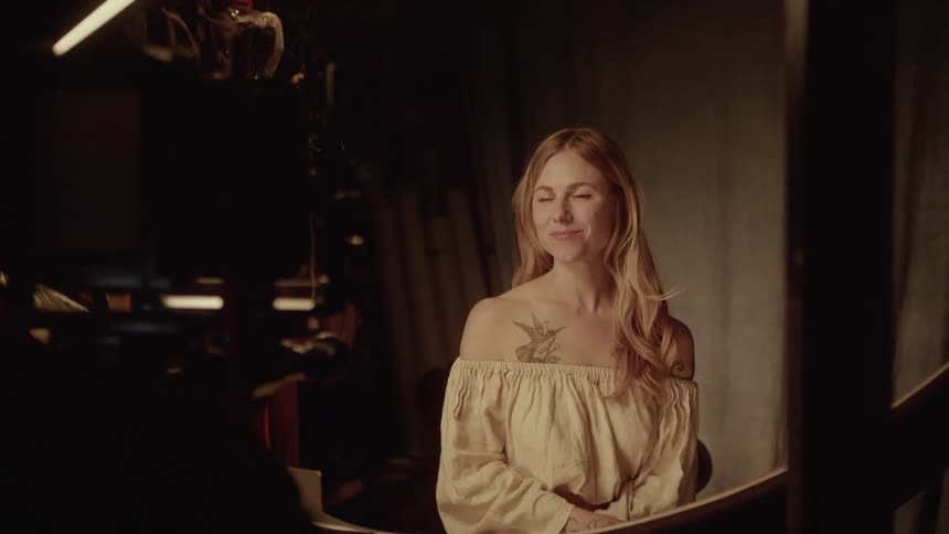 A blonde woman with a bird tattoo on her shoulder wearing a white cream blouse smiles while being filmed by a professional camera in a studio3