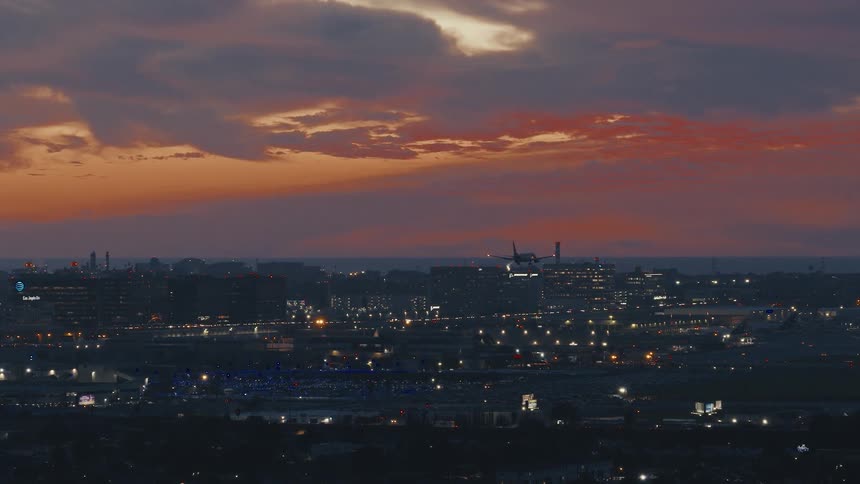 Lights glow in a city at dusk as traffic moves on the highways and an airplane comes in for a landing