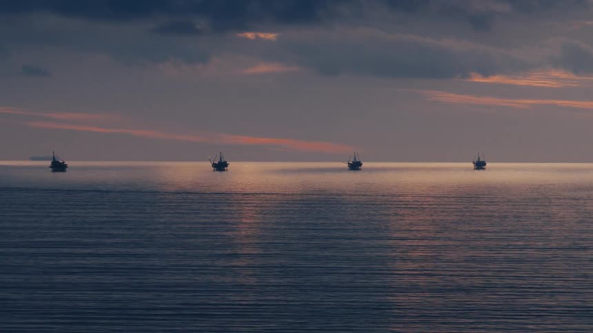 Calm ocean waters stretch toward offshore oil rigs silhouetted against a cloudy sunset horizon