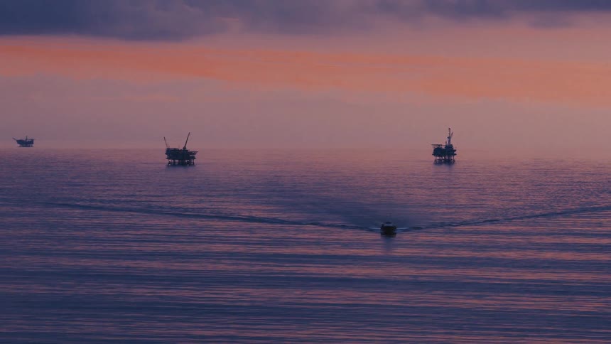 Ocean current forms a shallow angle near a structure while large buildings on stilts stand silhouetted against a bright reddish sky
