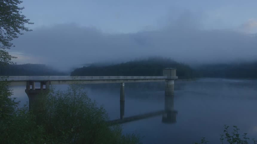 Bridge crossing a calm lake surrounded by fog with vegetation along the shore