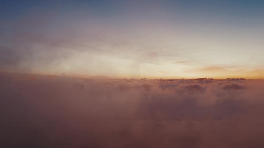 At dawn a soft golden glow spreads across the horizon as the sun rises above a vast sea of clouds tinting the mist with warm pinks and oranges beneath a calm fading blue sky