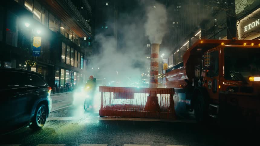A rainslicked city street at night traffic flows steadily as cars a utility truck and a lone cyclist all move past a glowing construction barrier and a steaming street vent with towering office buildings casting reflections across the wet pavement