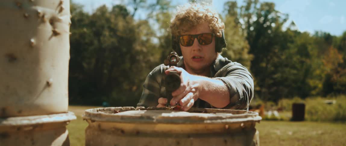 A man in a flannel shirt and sunglasses runs toward a barrel while holding a firearm he then crouches behind the barrel to steady his aim and points the weapon toward the camera