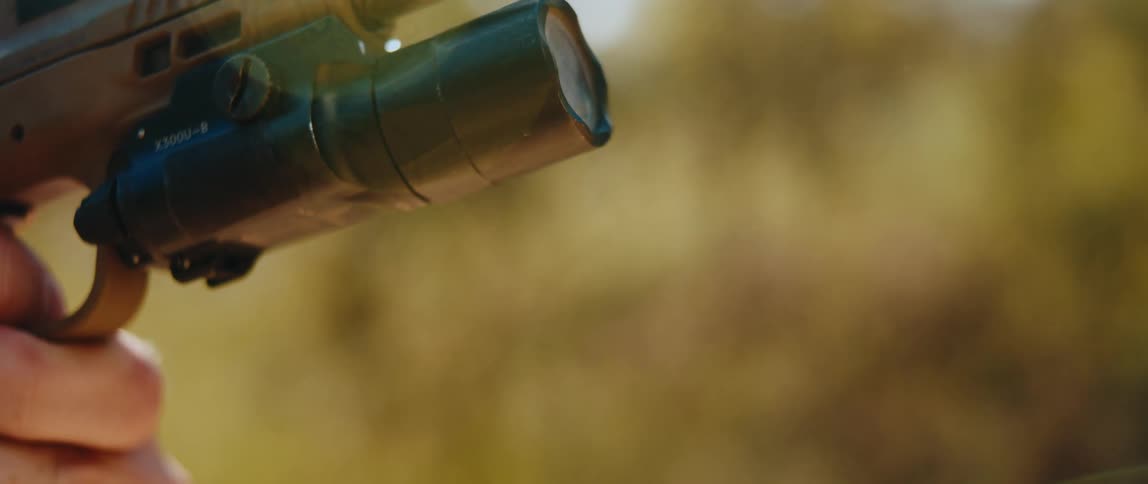 A tan semiautomatic pistol fires a round in slow motion showing the slide cycling back and the barrel tilting upward while a mounted tactical light remains attached to the frame against a blurred outdoor background