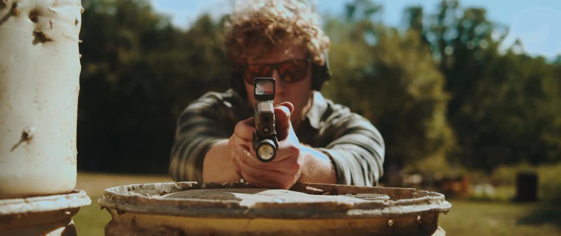 A man in a plaid shirt and safety gear draws a handgun and aims it directly at the camera he positions himself behind a weathered barrel in an outdoor setting focusing through a red dot sight