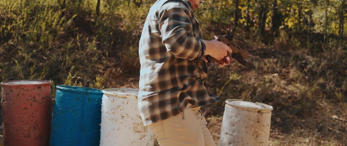 A man in a plaid shirt draws a handgun from an appendix holster clears his garment and extends the firearm into a twohanded firing stance at an outdoor range with colorful barrels in the background