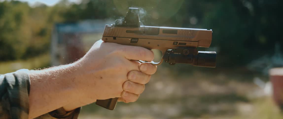 A closeup captures a handgun firing as a spent casing ejects upward from the slide with light smoke visible near the chamber a mounted optic and flashlight attached and a hand firmly gripping the firearm against an outdoor blurred background