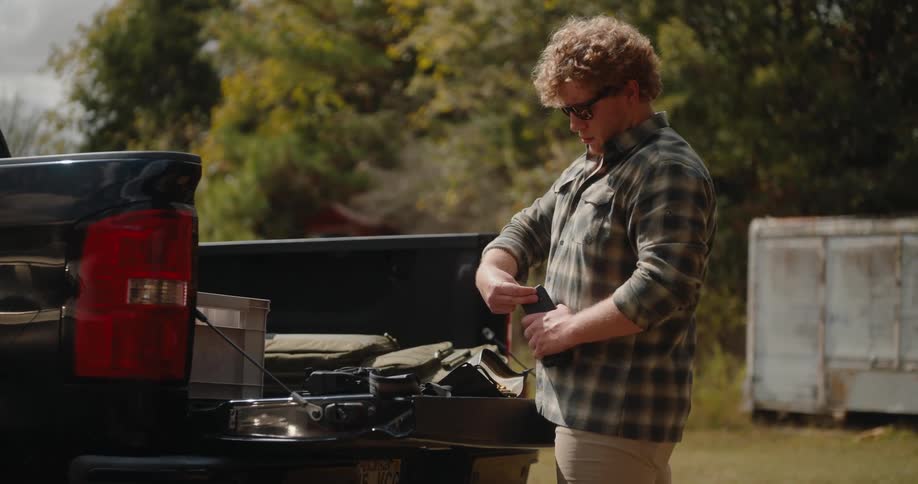 A man stands by a pickup truck loading ammunition in a rural outdoor area