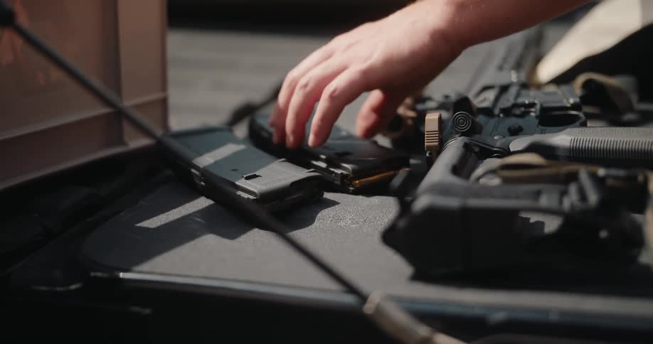 A hand reaches out to place a loaded magazine onto a flat surface next to a rifle the scene is set outdoors with several pieces of equipment resting in the back of a vehicle