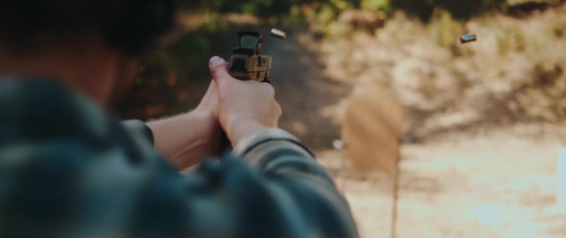 A man wearing a flannel shirt and ear protection fires a tan handgun equipped with a reddot sight at a cardboard target at an outdoor range with spent shell casings ejecting into the air after each shot