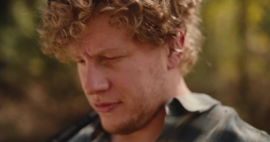 A curlyhaired man wearing a plaid shirt outdoors with his face partially turned downward looks forward with trees softly blurred in the background