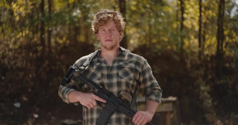 A man with curly hair stands outdoors and poses with his firearm