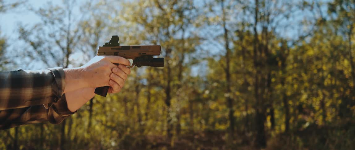A man draws a tan handgun from an appendix holster under his plaid shirt and fires several shots with visible muzzle flashes and brass casings ejecting into the air against a background of sunlit autumn trees and a white barrel