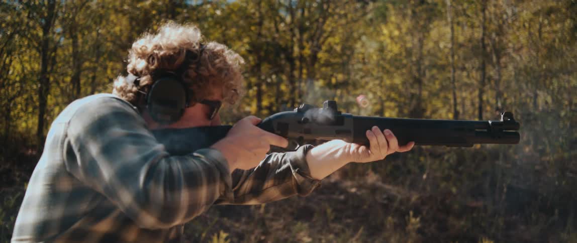 A man fires a shotgun in slow motion with an empty cartridge expelled as smoke rises from the barrel