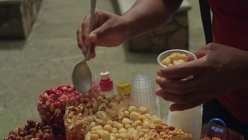 A close view shows a person using a silver spoon to scoop various nuts from large containers into a small plastic cup at an outdoor food stall