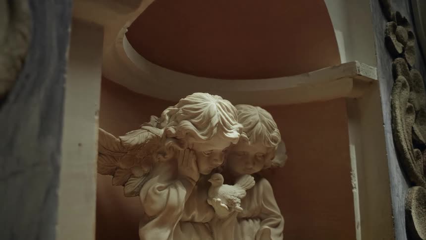 Two small angel statues are nestled together in a narrow architectural alcove one angel rests its head on its hand while both look down toward a small bird held in their center