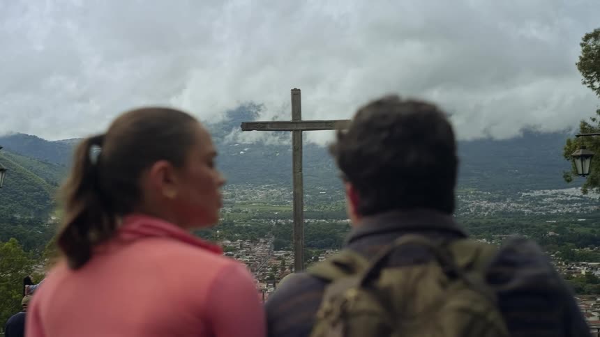 Couple looks at an elevated cross at a viewpoint overlooking a city and surrounding mountains