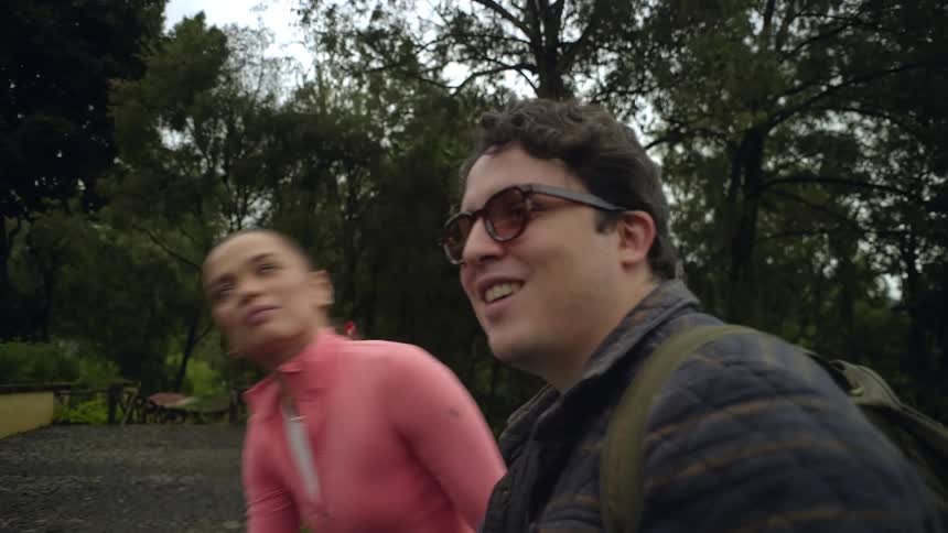 A man with glasses and a backpack looks toward a forest alongside a woman in a pink shirt before they walk together along a stone path with a wooden fence