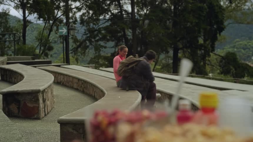 In the video a man carrying a backpack walks toward a woman who is sitting on a stone bench in a park setting he sits down beside her and begins to eat something from a container as they start a conversation
