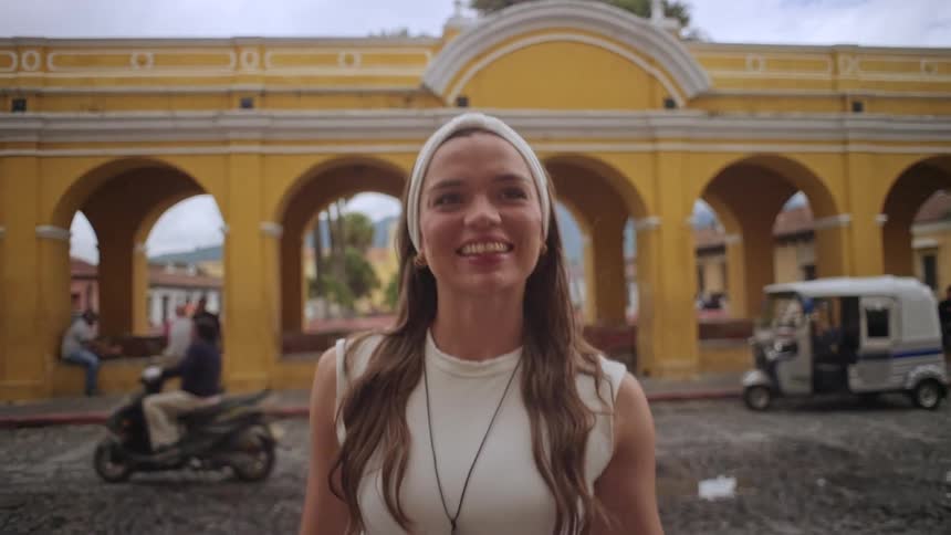 A woman poses with a bright smile in front of a yellow arched building as a motorcycle passes by in the background