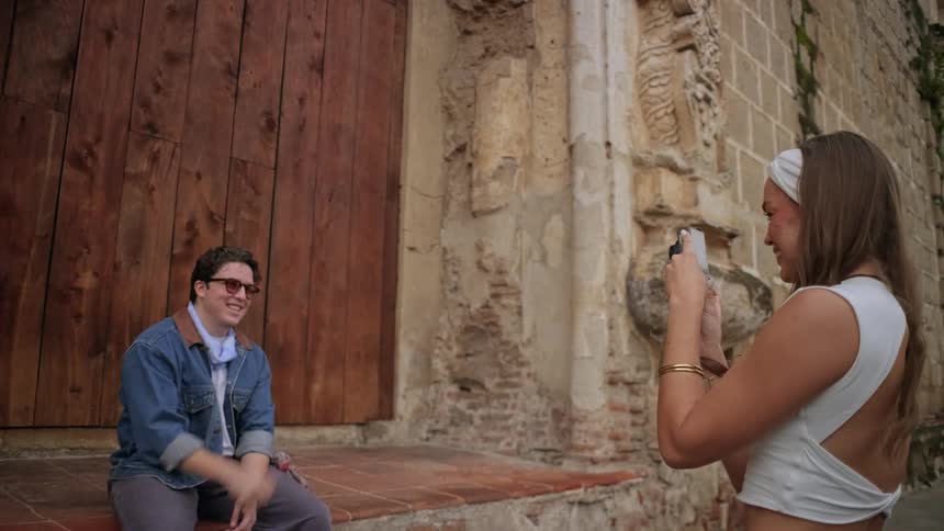 A young woman in a white dress taking a photo of a young man in a jean jacket and sunglasses posing while seated in front of a door at midday