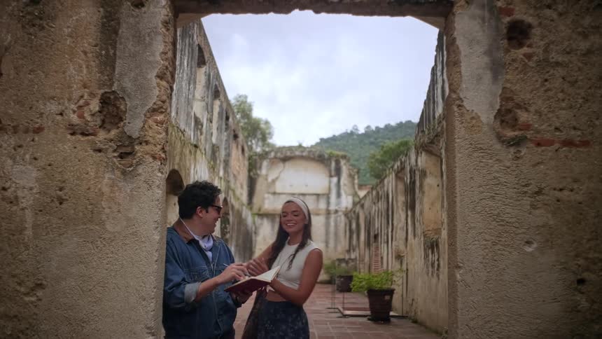A man in a denim jacket stands in stone ruins writing in a notebook when a woman in a white headband approaches him causing them both to laugh and look at the book together against a backdrop of weathered walls and a cloudy sky