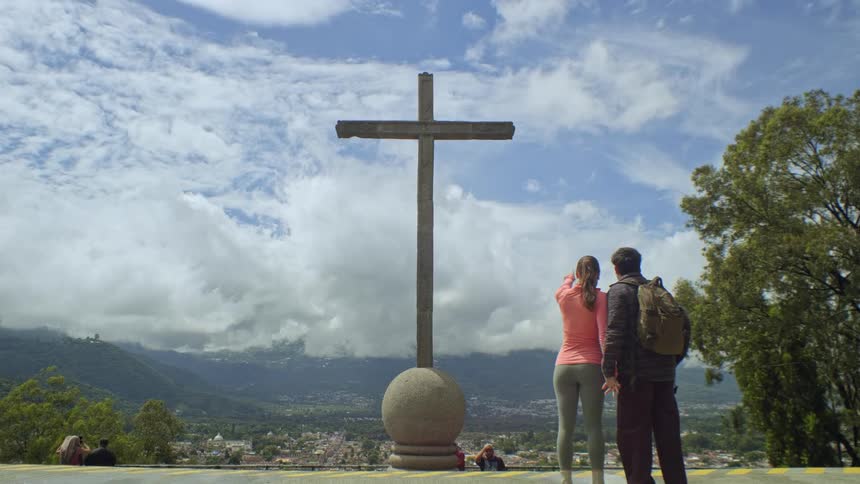 Couple at a viewpoint pointing toward an urban and mountainous landscape with a large wooden cross in the foreground