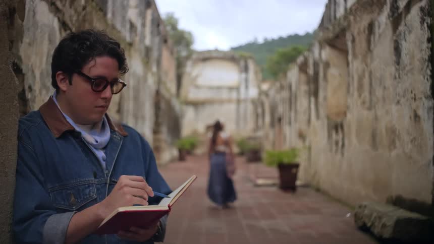 A young man in a jean jacket and sunglasses writing in a notebook with a pen while a woman walks in the background through a roofless ruined building at midday