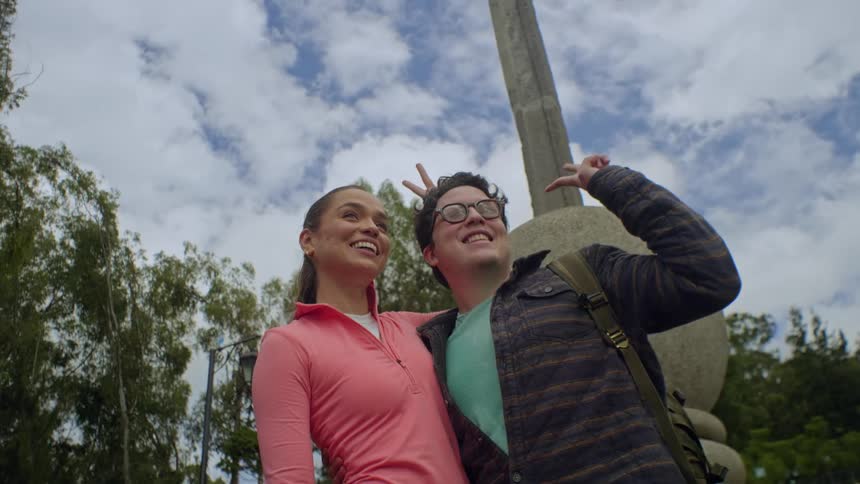 Couple poses smiling in front of an outdoor monument under a partly cloudy sky