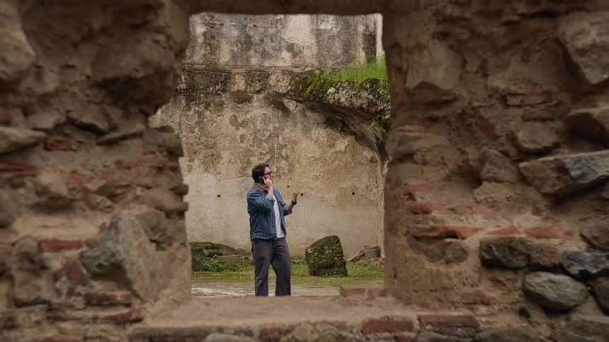 Man talks on a phone in a courtyard of historic ruins framed by a stone wall opening