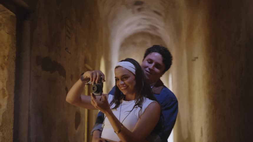Woman holding a camera while a man embraces her from behind in a narrow corridor with old walls
