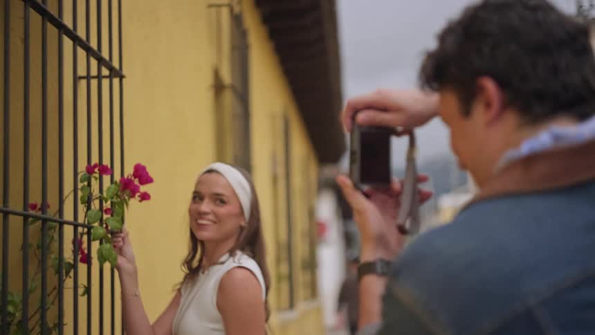 Woman examining flowers on a barred window while a man photographs the scene on an urban street