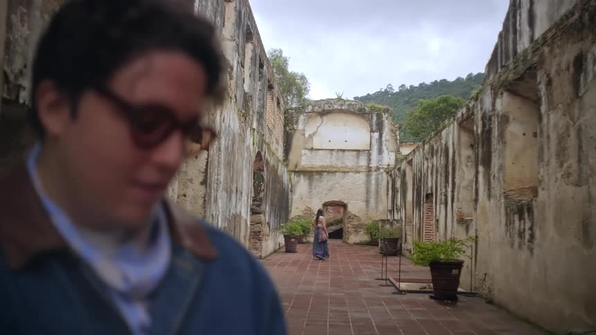 A man with glasses stands in the foreground of a large stone ruin while a woman walks through a central doorway in the background under a cloudy sky