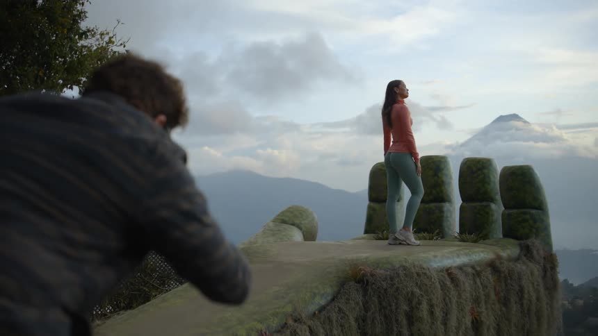 A photographer captures a woman posing on a mountain overlook with cloudy peaks in the background
