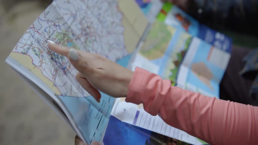 A person in a pink longsleeve shirt points their finger at specific locations on a paper map while holding it with another person outdoors