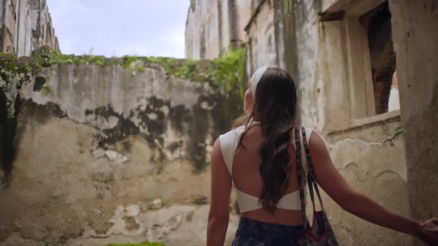 A woman with long brown hair walks through the stone ruins of an ancient roofless buildingshe gazes up at the weathered walls and greenery while carrying a patterned bag over her shoulder
