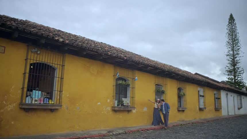 Couple walks along a cobblestone street beside a yellow building with barred windows